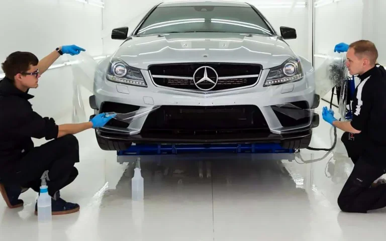 Un detailer profesional y su compañero trabajan el frontal de un Mercedes plateado en una cabina blanca